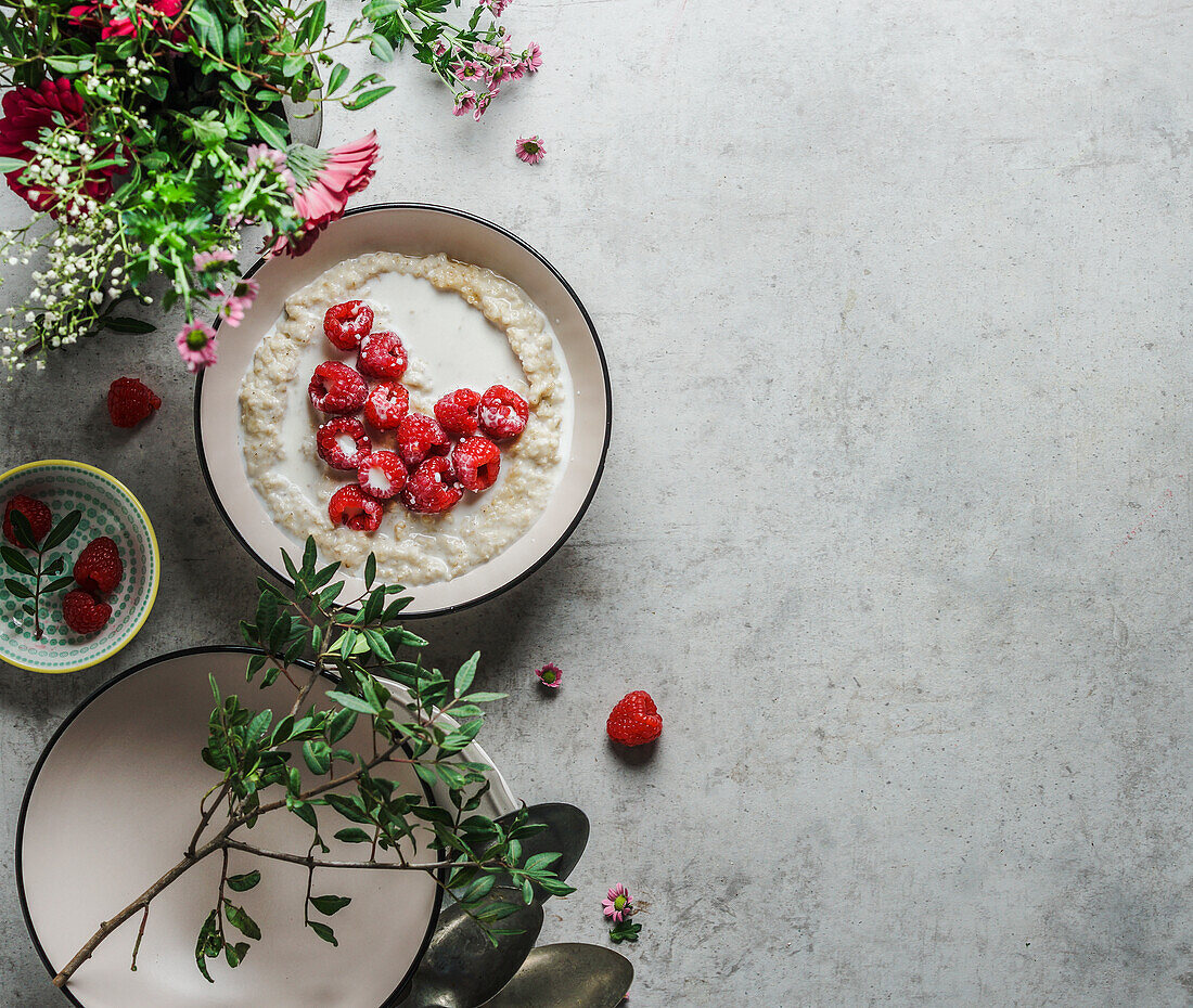 Romantisches Frühstück mit Porridge und einem Herz aus Himbeeren auf einem grauen Küchentisch mit Blumen und Schüsseln. Vegane Frühstücksidee mit nahrhaften Zutaten. Draufsicht mit Kopierbereich