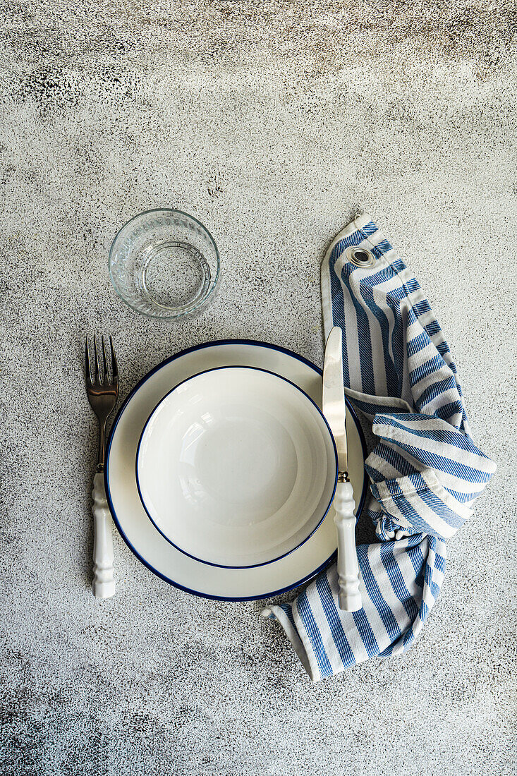 Top view of minimalistic rustic table setting with white plates, cutlery, glass and striped napkin on gray surface