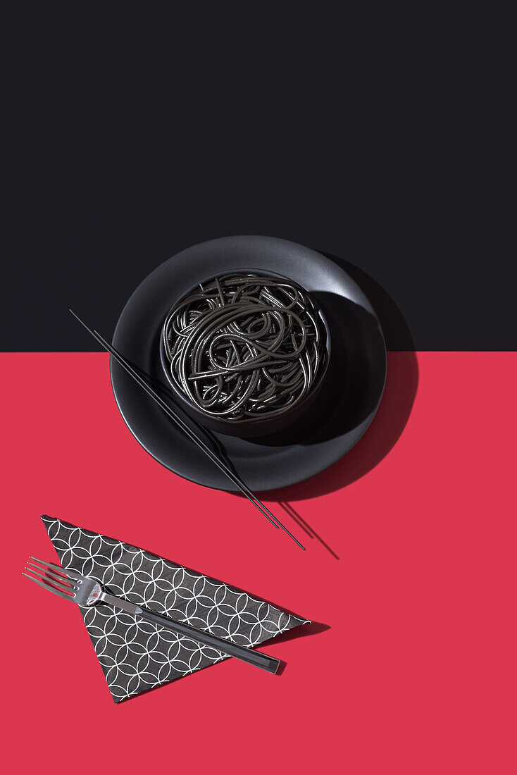 Draufsicht auf schwarze Spaghetti in einer Schüssel mit Stäbchen auf schwarzem und rotem Hintergrund neben Serviette und Gabel