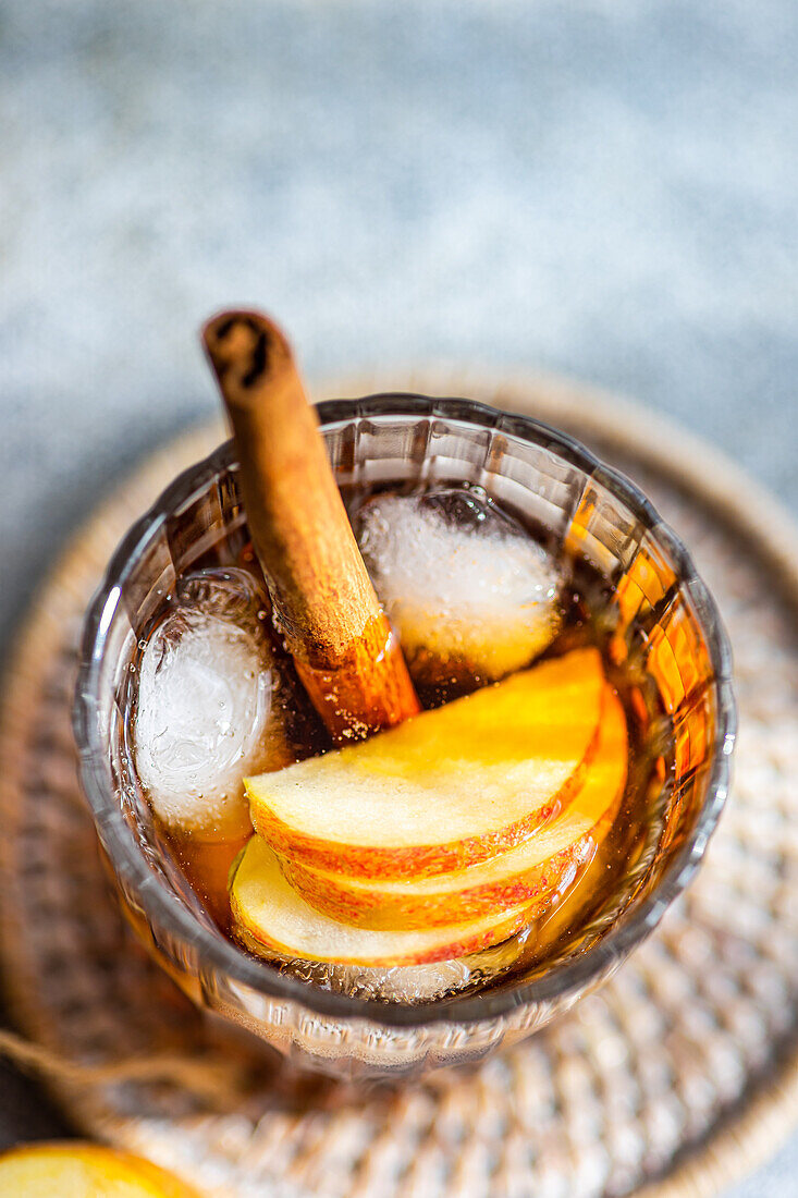 Draufsicht auf einen frischen Apfelweincocktail, garniert mit Zimtstangen, Sternanis und Apfelscheiben in einem mit Eiswürfeln gefüllten Kristallglas auf einem strukturierten Hintergrund