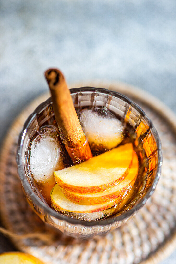 Top view of fresh apple cider cocktail garnished with cinnamon sticks, star anise, and apple slices in a crystal glass filled with ice cubes on a textured background