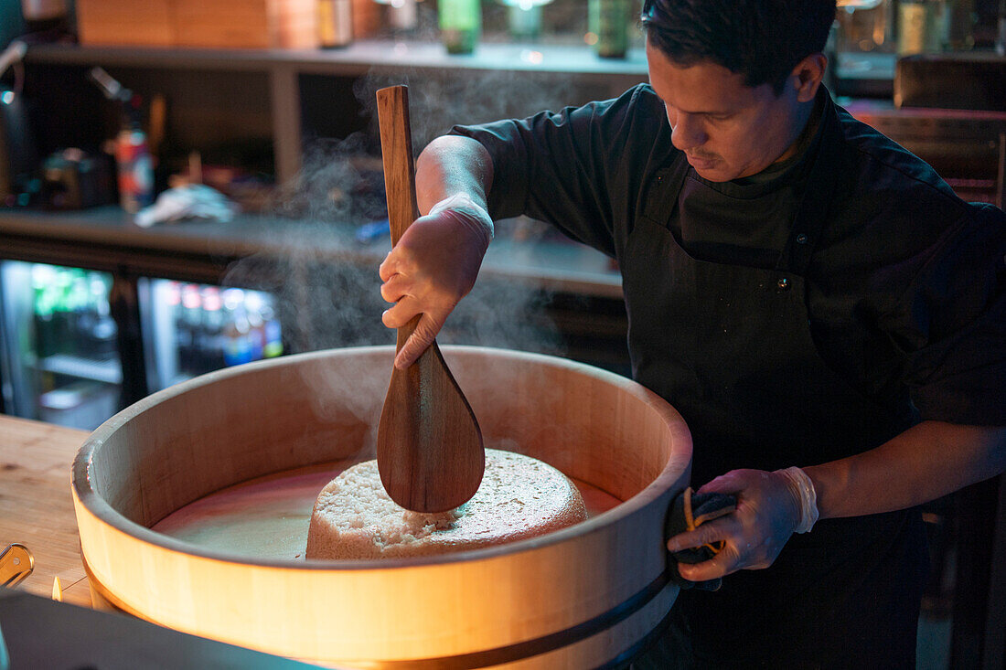 An expert sushi chef carefully mixes sushi rice in a wooden tub known as a hangiri at a sushi restaurant.