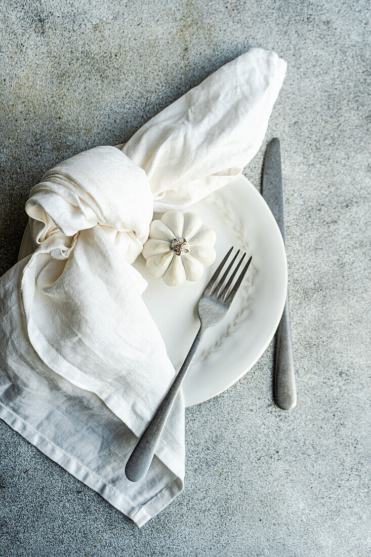 Draufsicht auf weißen Keramikteller mit Zierkürbissen mit silberner Gabel und Messer, eingewickelt in eine weiße Serviette, platziert auf einer strukturierten grauen Betonfläche