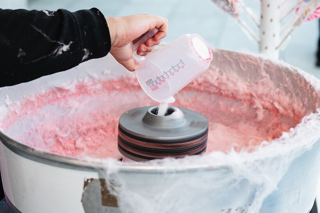 Close-up of a cropped unrecognizable hand pouring sugar into a cotton candy machine with pink candy floss forming inside