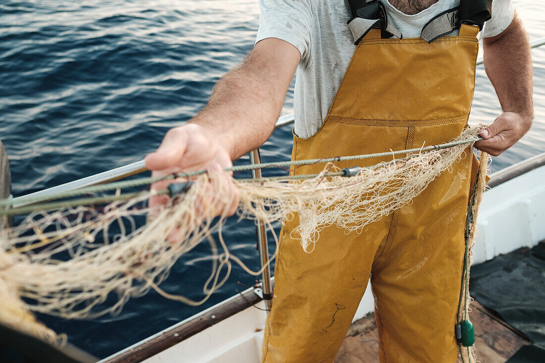 Von oben abgeschnittener unkenntlicher männlicher Fischer in Uniform, der mit einem Netz auf Fischfang geht, während er auf einem Schoner in Soller nahe der Baleareninsel Mallorca arbeitet