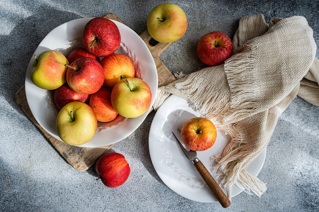 Sammlung reifer, farbenfroher Äpfel auf einem rustikalen Holzbrett, mit weißem Teller, Gabel und drapiertem Stoff, der eine heitere Küchenatmosphäre schafft, von oben