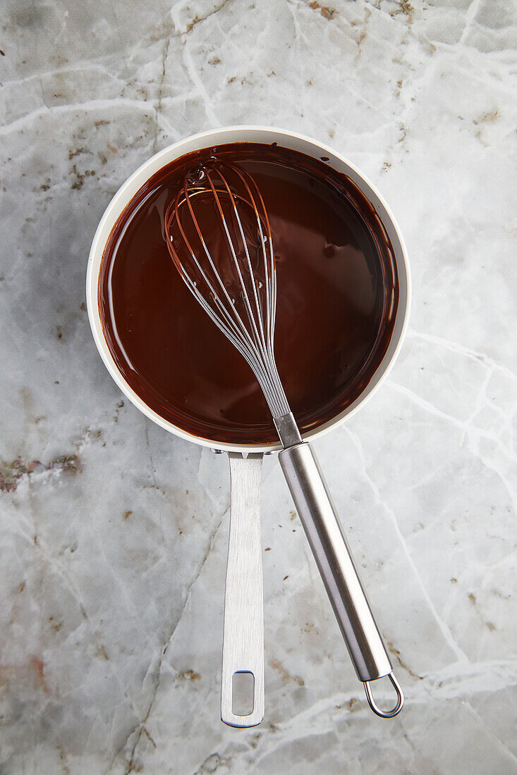 Draufsicht auf geschmolzene heiße Milchschokolade in einem Metalltopf mit Schneebesen, der auf einer Marmortheke für die Zubereitung von Gebäck steht