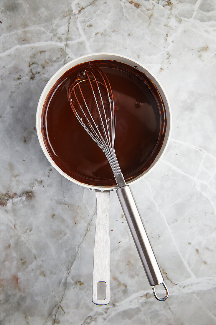 Top view of melted hot milk chocolate in metal saucepan with whisk placed on marble counter for pastry cooking