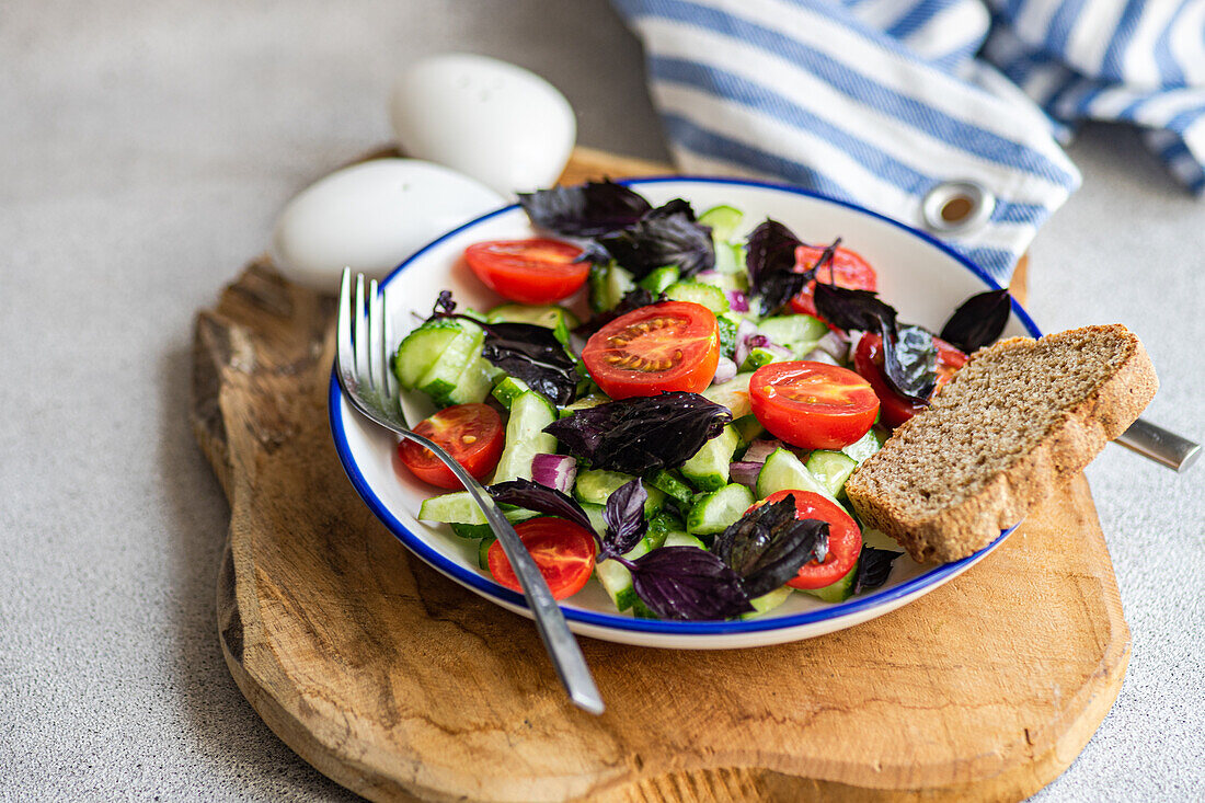 Hochformatiger Gemüsesalat der Saison mit Bio-Gurke, Tomate, Zwiebel und roten Basilikumblättern, serviert auf einem Teller mit gestreifter Serviette vor grauem Hintergrund