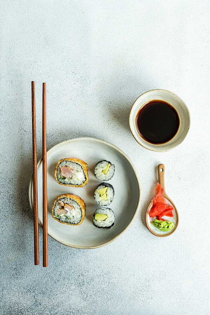 Blick von oben auf ein japanisches Essensset mit Avocado-Maki-Rolle und gebratenem Sushi mit Sojasauce und Ingwer auf einem weißen Betontisch