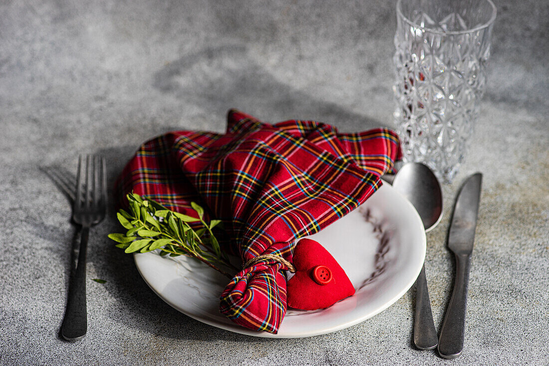 Top view of ceramic plate with cutlery, tartan fabric napkin with heart shaped decor placed on concrete surface at kitchen table for meal during Valentine's day celebration