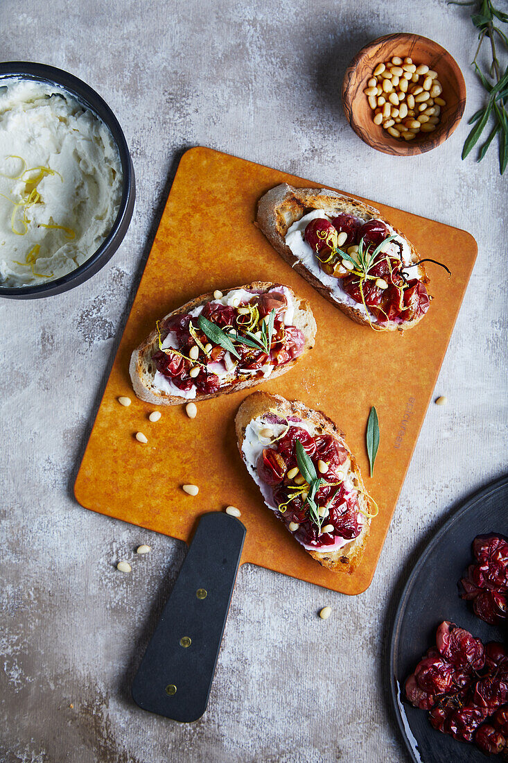 Top view of appetizing ricotta toast with roasted grapes and lemon zest served with tarragon and pine nuts placed on crusty bread on wooden board on gray table