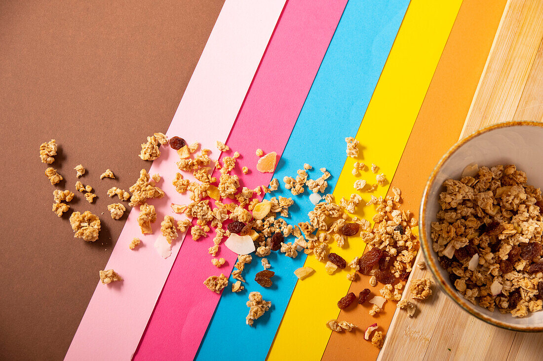 Top view of scattered granola with muesli in bowl and on surface with multicolored ribbons placed on wooden table in daylight