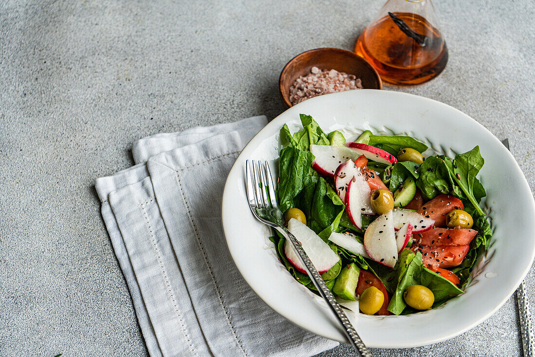 Freshly prepared vegetable salad featuring organic spinach leaves, ripe tomatoes, crisp radishes, and green olives beautifully presented in a white bowl with a fork set against a muted backdrop