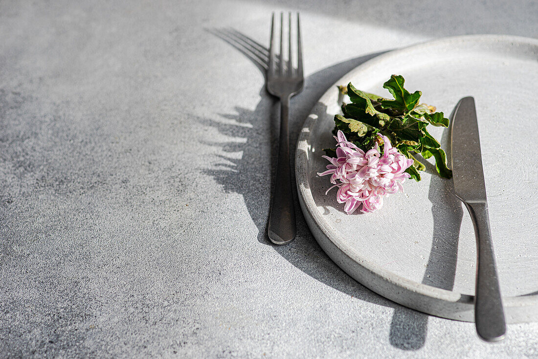 A minimalistic table setting featuring a white plate with a pink flower and green leaves, alongside elegant cutlery on a textured surface.