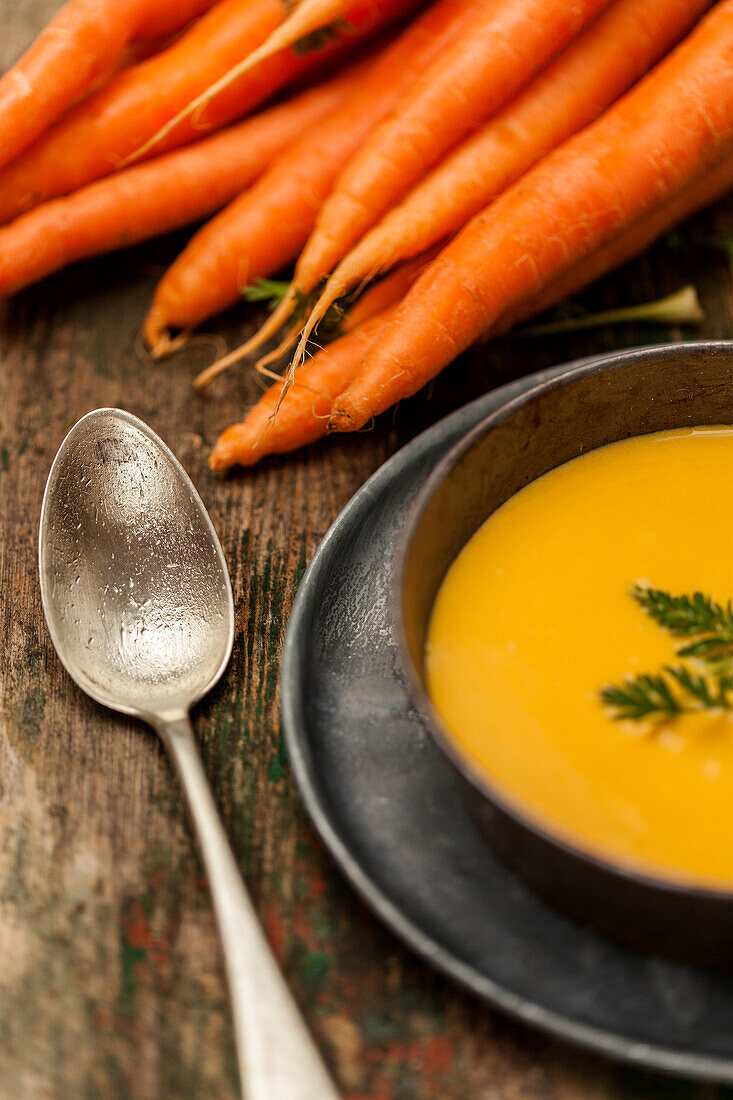 Frische Karotten neben einer Schüssel mit cremiger hausgemachter Karottensuppe, garniert mit Kräutern, präsentiert auf einem rustikalen Holztisch