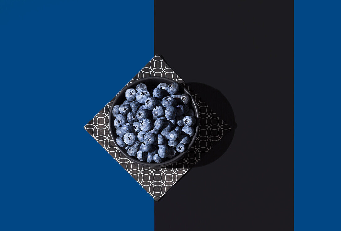Top view of bowl of juicy blueberries on a geometric patterned napkin, set against a contrasting blue and black diagonal background