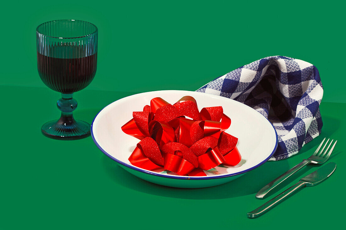 Generative AI image featuring a plate filled with glittering red ribbons, a checkered napkin, cutlery, and a dark wine glass on a green background, symbolizing a quirky take on Valentine's Day romance.