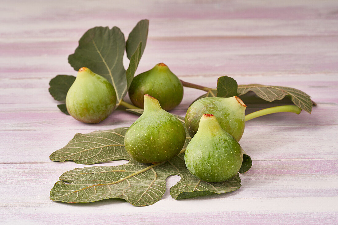 Ripe green figs accompanied by their leaves laid out on a pink wooden surface, conveying a natural and healthy look.