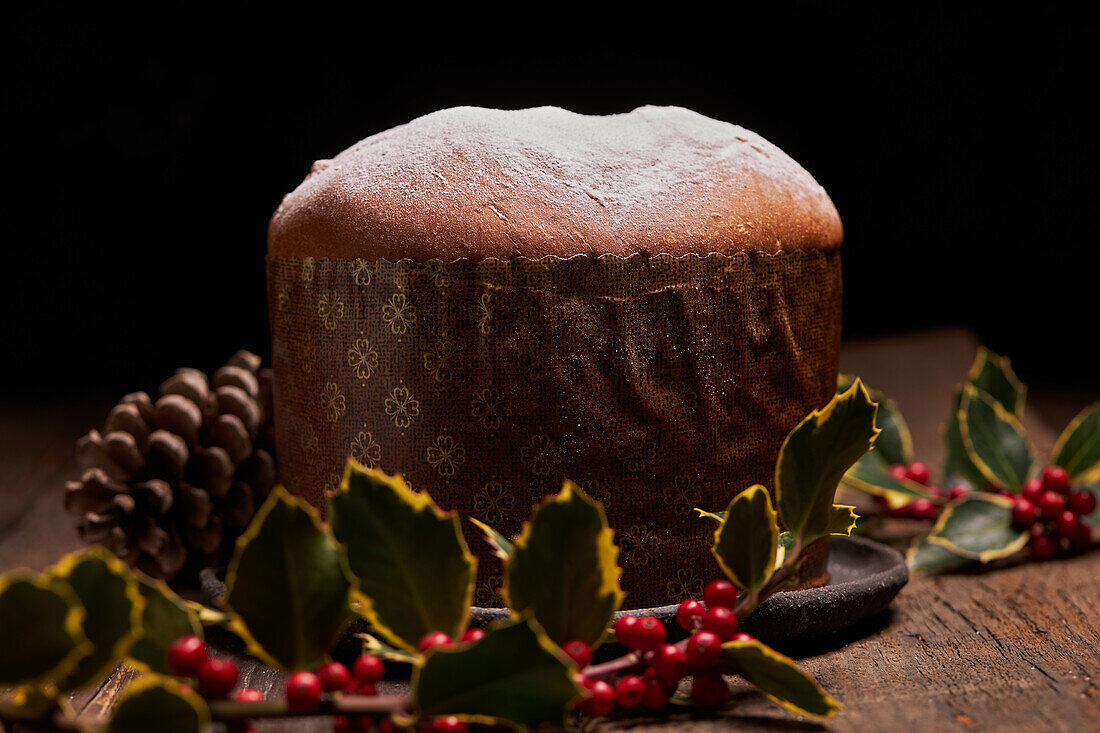 A beautifully presented traditional panettone with a light dusting of icing sugar, set against a backdrop of festive decor including holly berries and pinecones