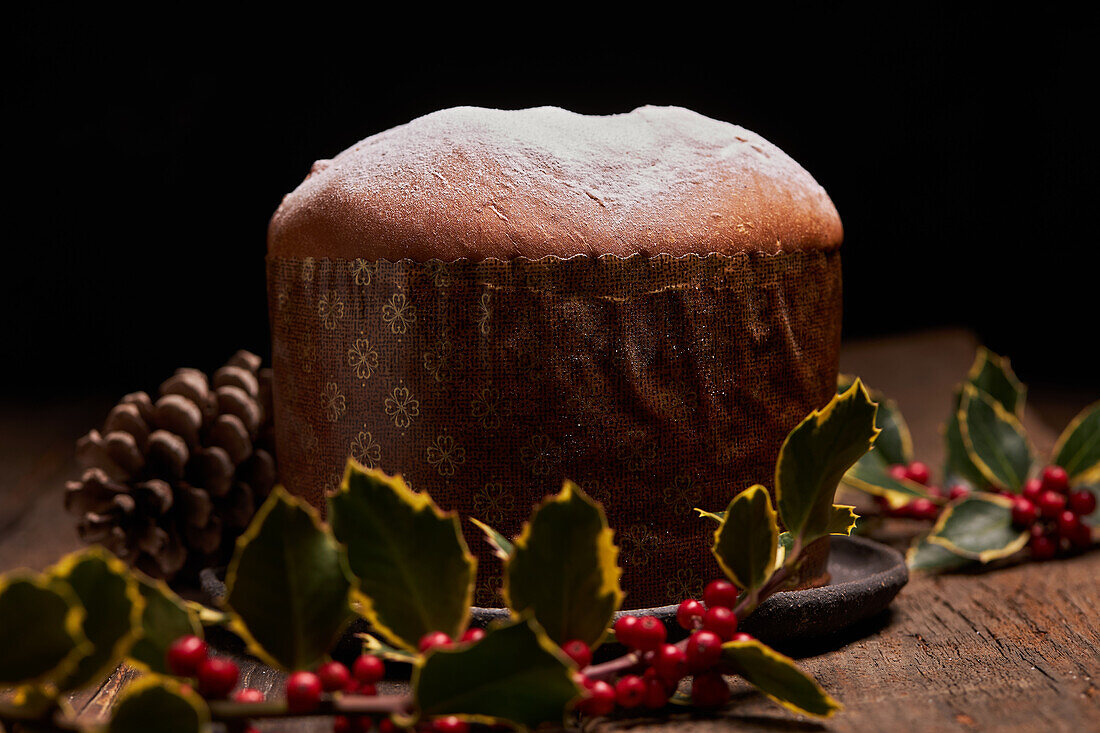 Ein wunderschön präsentierter traditioneller Panettone mit einer leichten Schicht Puderzucker, vor einem Hintergrund mit festlicher Dekoration, darunter Stechpalmenbeeren und Tannenzapfen