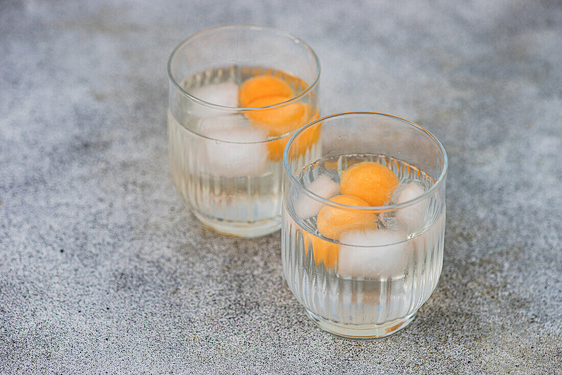 Blick von oben auf ein Paar transparente Gläser, gefüllt mit Melonenkugeln in einem eisgekühlten Getränk, die auf einer grauen Fläche im Tageslicht stehen