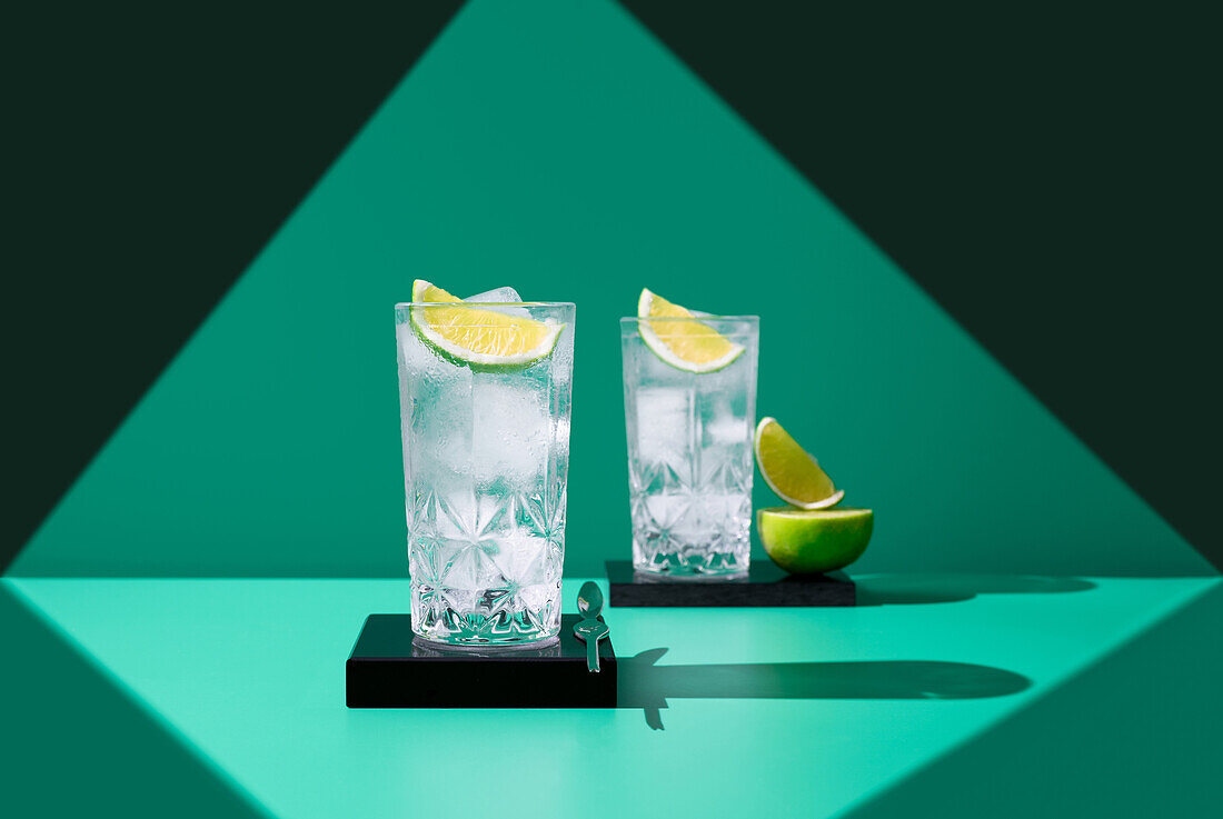 Two glasses of gin tonic with lime slices cast interesting shadows on a green background, highlighting a sense of refreshment.