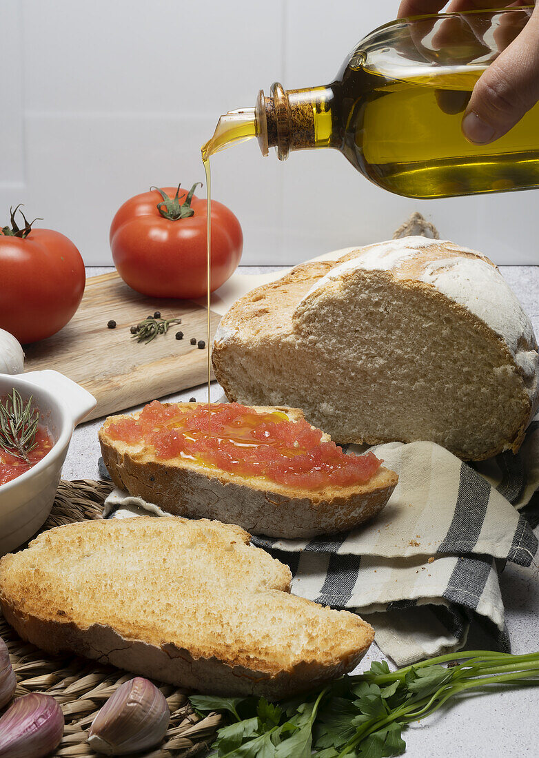 Die Hand einer anonymen Person gießt Olivenöl aus einer Flasche auf ein Vollkornbrot mit Tomatenaufstrich zwischen ungeschältem Knoblauch und einem Schneidebrett auf dem Tisch
