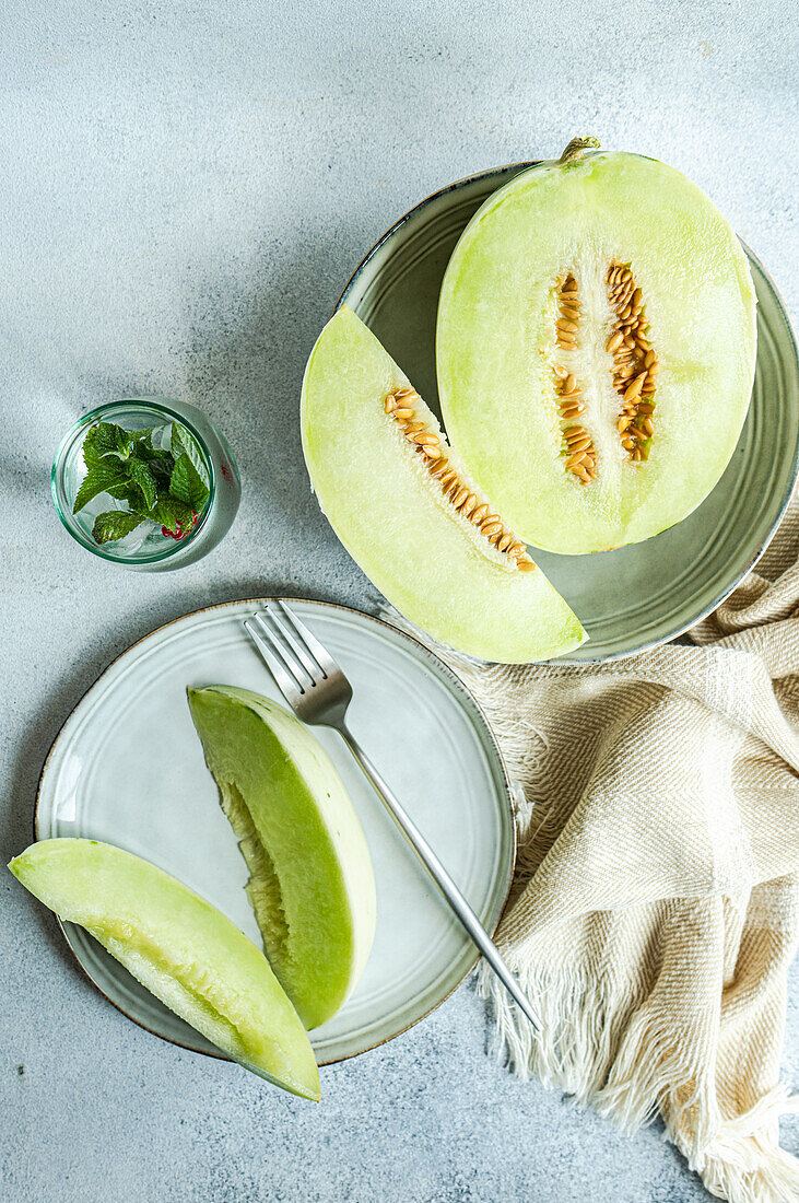 Blick von oben auf eine halbierte grüne Melone in einer Schale und auf einem Teller mit einer Limonade auf einem Betonhintergrund