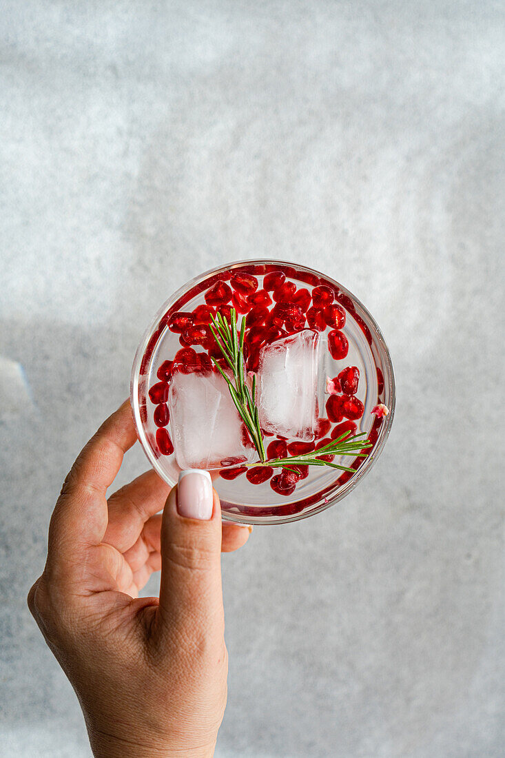 From above anonymous of hand holding a gin tonic cocktail with pomegranate seeds and rosemary over a grey textured background