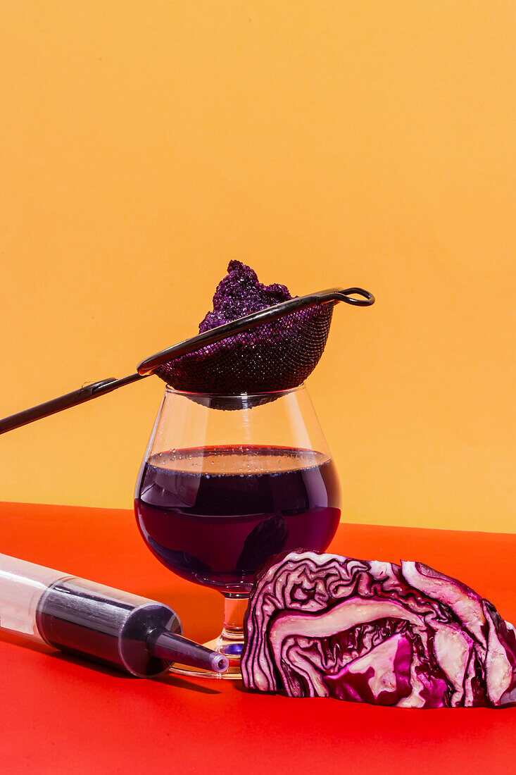 Composition of glass with strainer and syringe filled with homemade juice of red cabbage against bright background