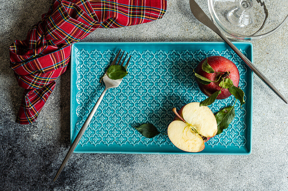 Top view of whole fresh red ripened apple with green leaves and cut half piece placed on blue tray near checkered napkin water glass on gray surface in daylight