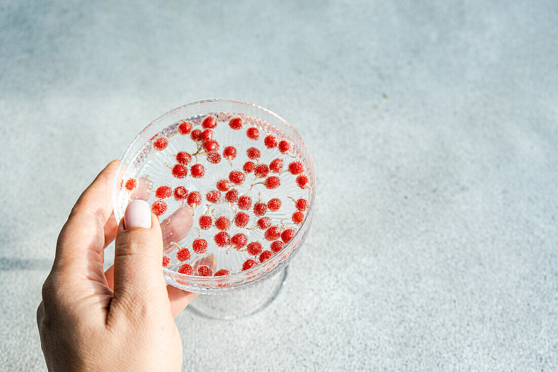 Eine zarte Hand präsentiert eine transparente Untertasse, die mit kleinen roten Beeren gefüllt ist und einen Schatten auf eine strukturierte Oberfläche wirft