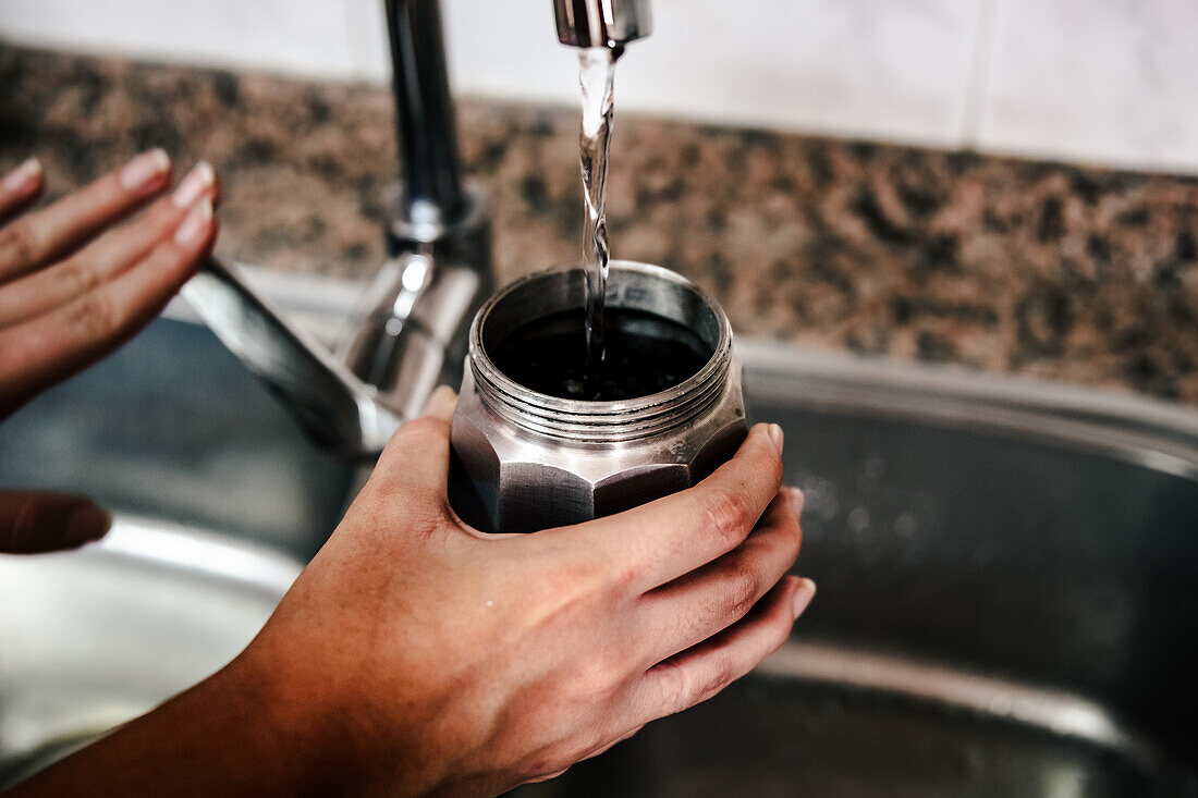 Nahaufnahme einer anonymen Hand, die eine italienische Mokkakanne mit Wasser aus einem Küchenhahn füllt, um die ersten Schritte der Zubereitung von traditionellem, selbst gebrühtem Kaffee hervorzuheben