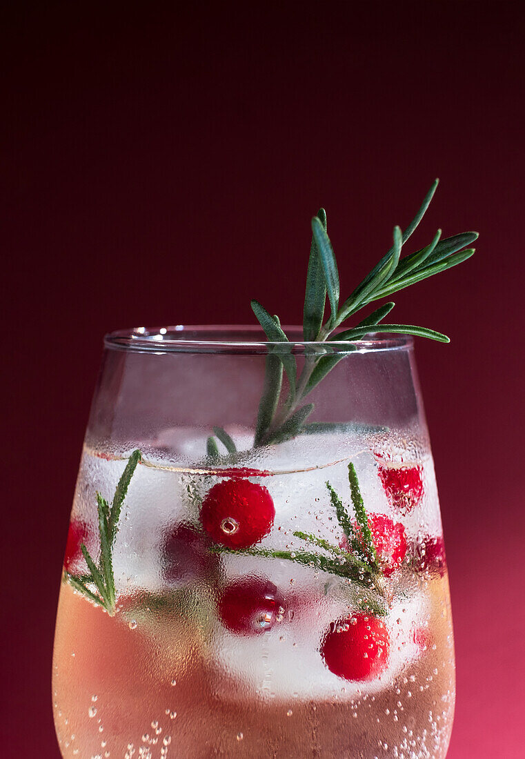 A sparkling Rosemary winter cocktail garnished with cranberries and a sprig of fresh rosemary, presented against a deep red background.