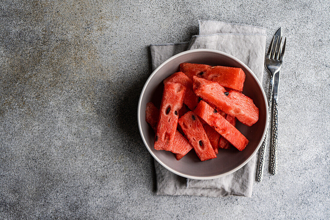 From above cut pieces of watermelon in bowl on tablecloth with cutlery on the side on concrete background