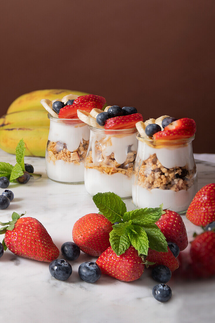 Yogurt whit fresh strawberries and blueberries with sliced banana placed in cups and on white marble table bright room