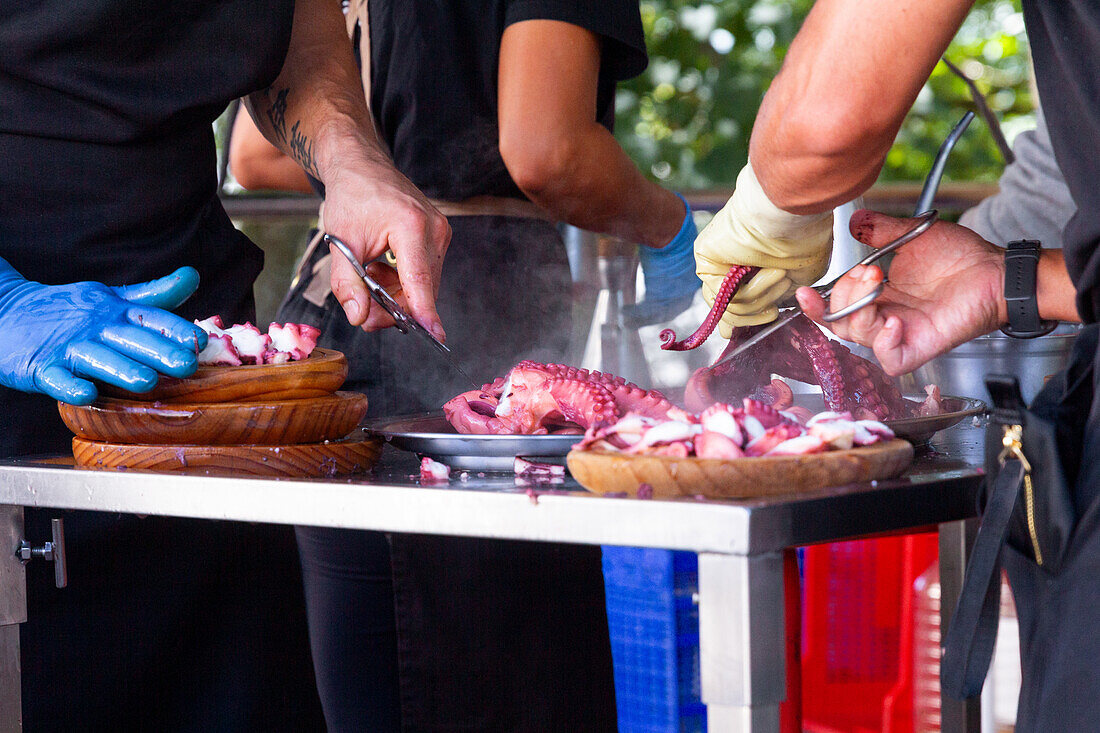 Anonymous chefs in gloves expertly prepare and cut octopus on a metal table, with wooden bowls and tools, set against an outdoor backdrop