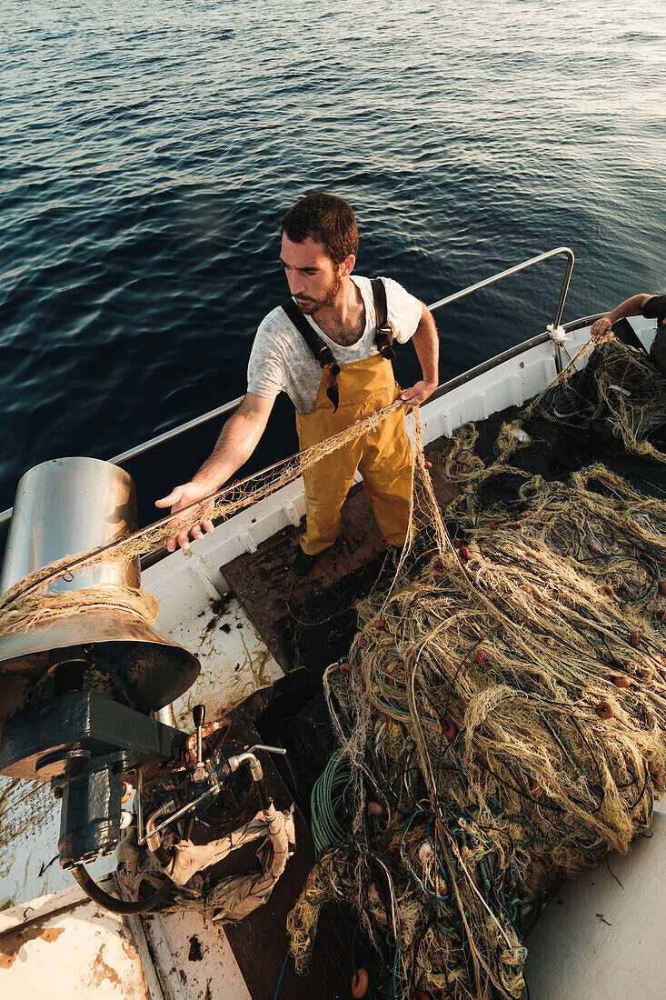 Von oben fokussierter bärtiger männlicher Fischer in Uniform, der mit einem Netz auf Fischfang geht, während er auf einem Schoner in Soller nahe der Baleareninsel Mallorca arbeitet