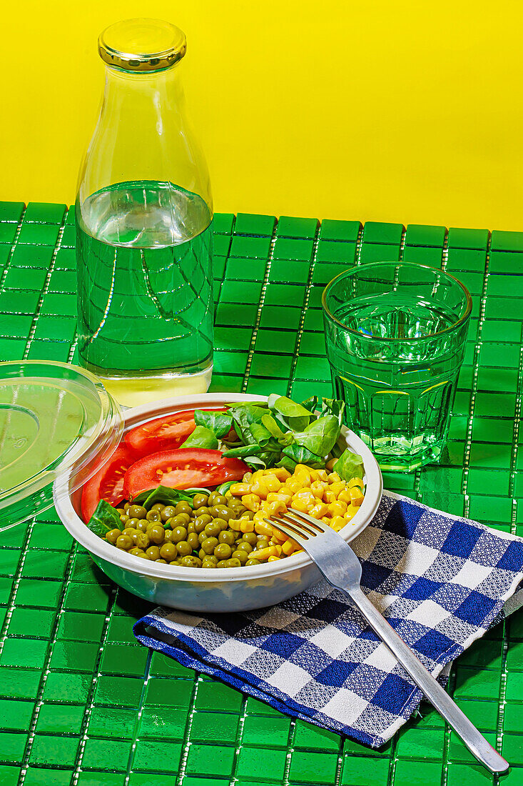 High angle of salad bowl with slices of tomato, spinach leaves, corn kernels and peas placed on napkin on green surface with fork near glass and bottle of water against yellow wall