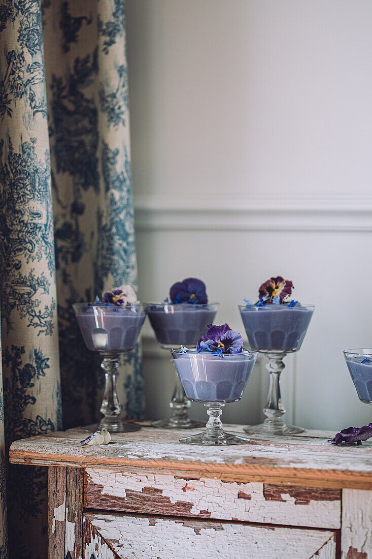 Verschiedene Gläser mit schmackhaftem, blauem Spirulina-Pudding, dekoriert mit Blumen, auf einem schäbigen Holzschrank