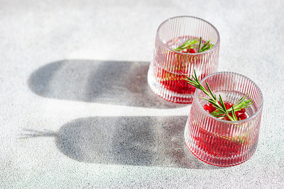 Blick von oben auf sonnenbeschienene Gin-Tonic-Cocktails mit Granatapfel und Rosmarin in strukturierten Gläsern auf einer hellen Fläche