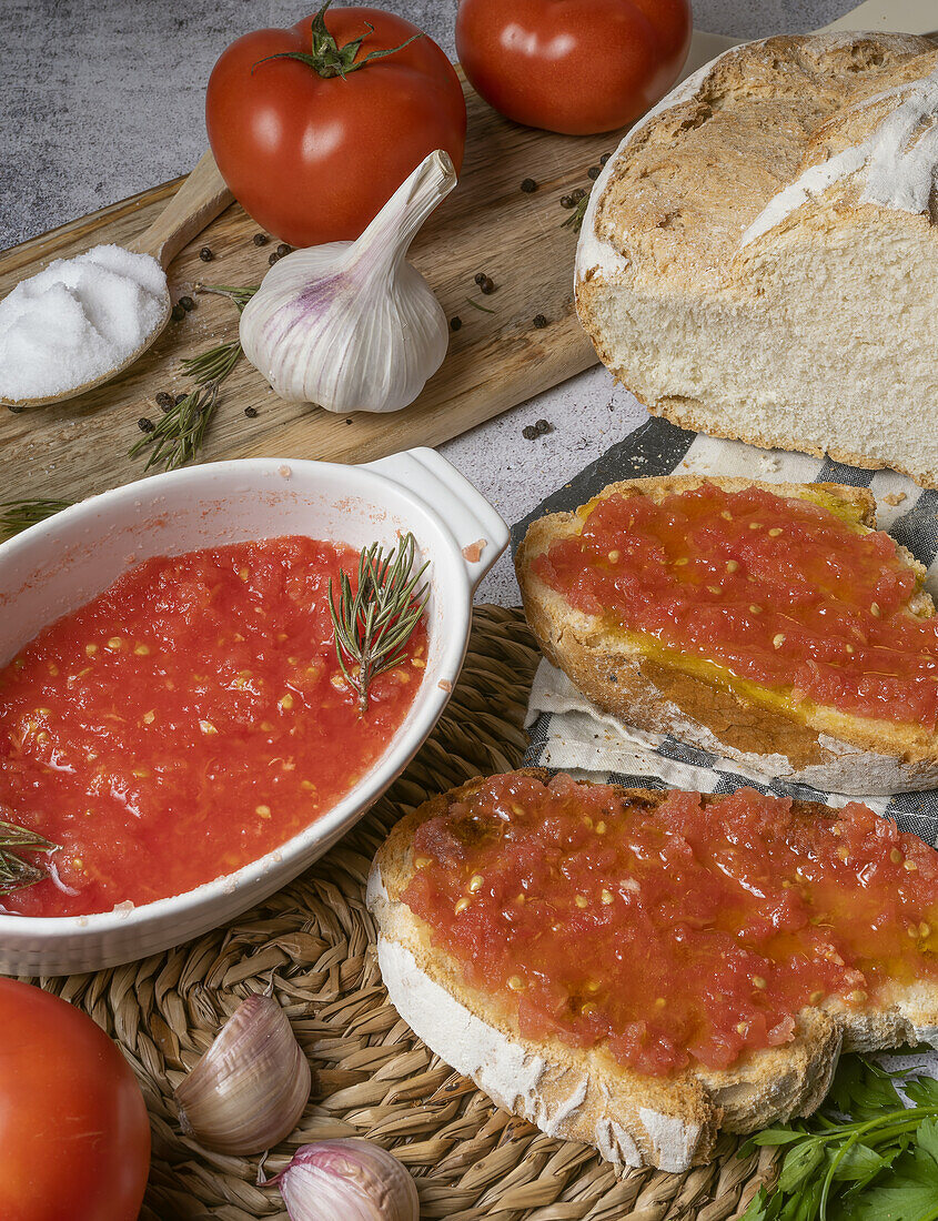 From above of pieces of bread with tomato spread placed on table near bowl with tomato spread, chopping board, garlic and spoon with salt