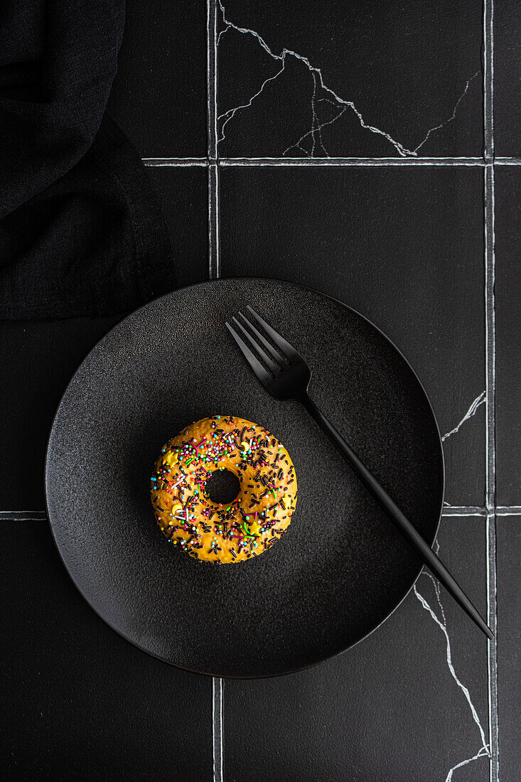 Top view of sweet banana donut with colorful sprinkles placed on black plate with fork against dark surface