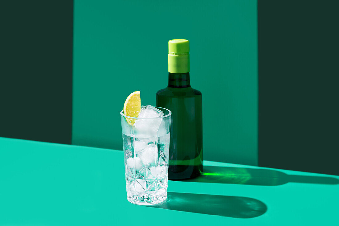 A refreshing gin tonic with ice cubes and a lime wedge, beside a bottle of gin, set against a green background with shadows.