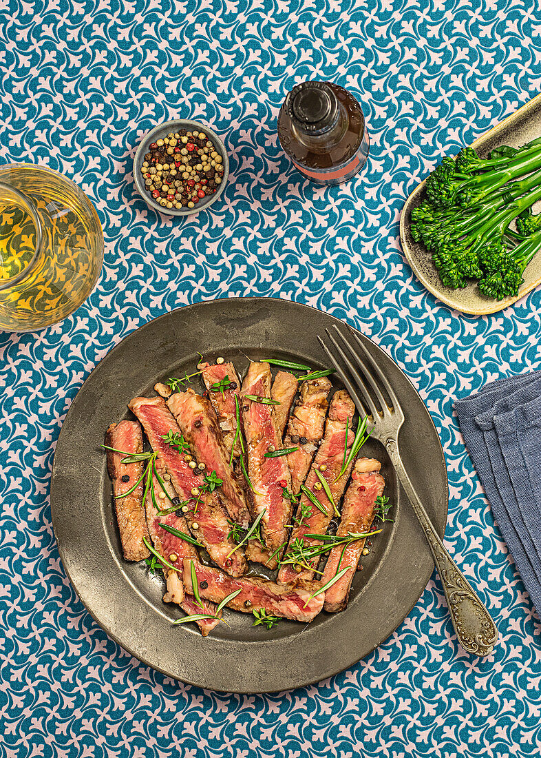 Von oben köstliche Roastbeef-Tagliatta auf einem Teller neben einer Schüssel mit Brokkoli und Kräutern auf einem gemusterten Tischtuch serviert