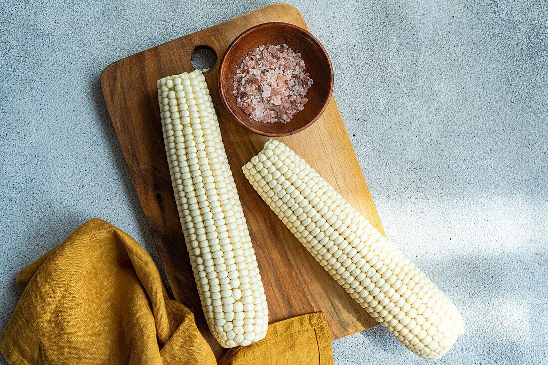 Overhead wooden chopping board with ripe corncobs placed on concrete table in kitchen with bowl with sea salt and napkin