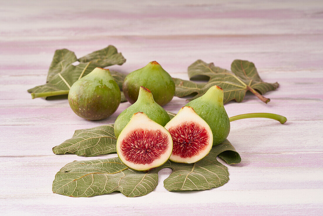 Green figs and their leaves are artfully arranged on a pale wooden surface, with one fig cut open to display the red interior.
