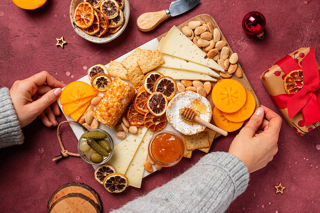 Hands arranging a festive cheese board with various cheeses, nuts, dried fruits, and holiday decorations on a textured surface.