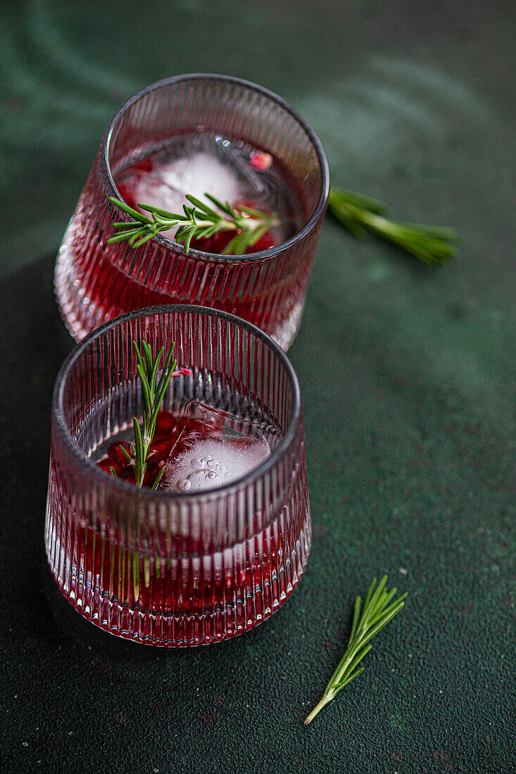Top view of two glasses of gin tonic cocktail with pomegranate seeds and a sprig of rosemary, on a dark green textured surface with a single rosemary sprig beside